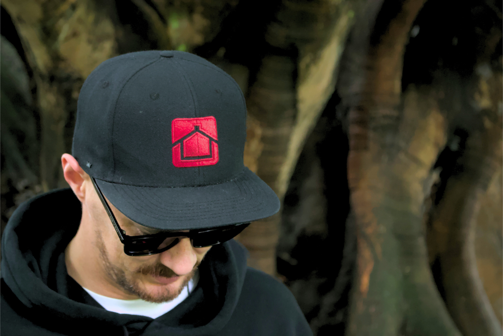 Person wearing a black cap with a red logo and sunglasses, standing against a blurred natural background.