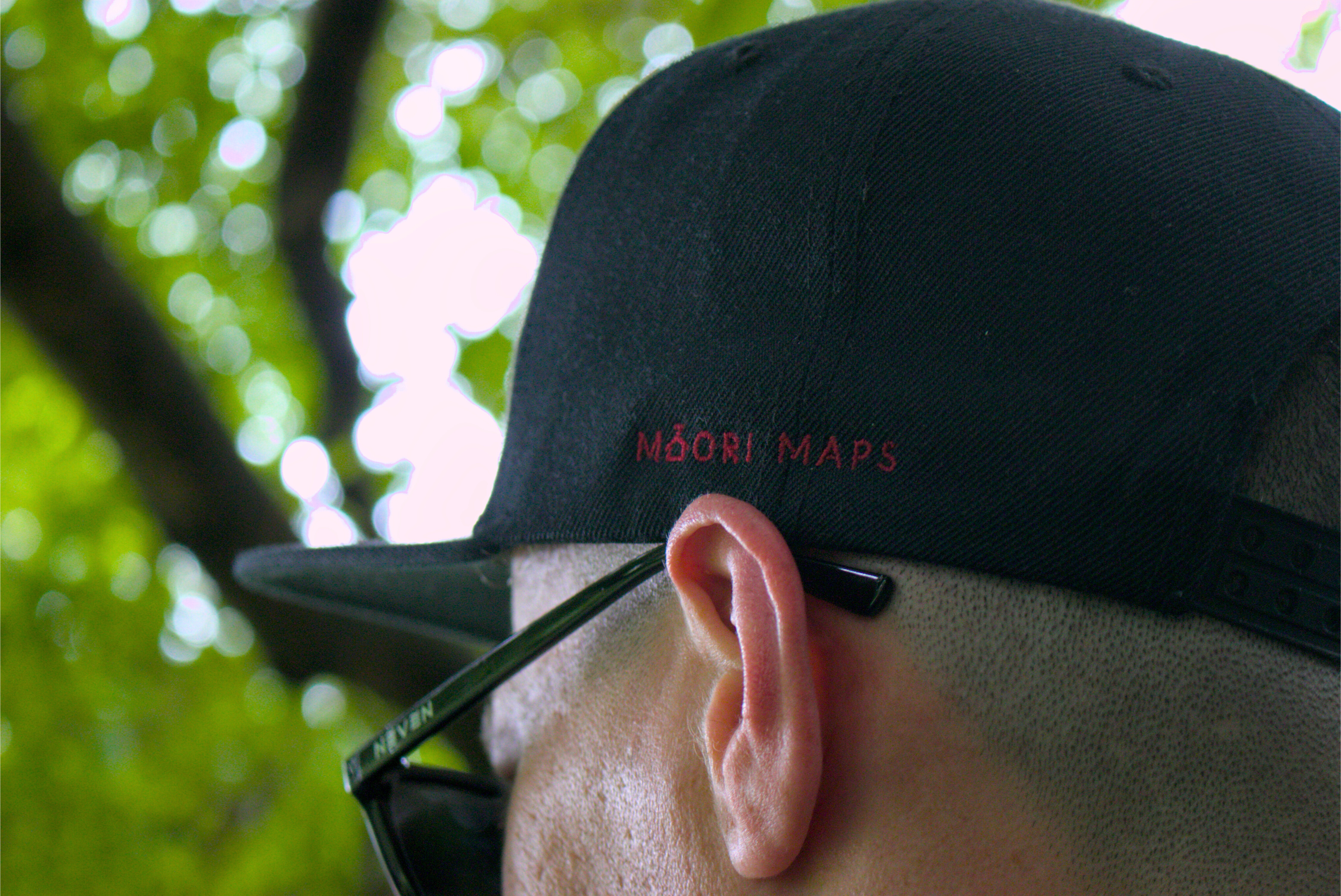 Person wearing a black cap with 'Māori Maps' text, sunglasses on head, blurred green background