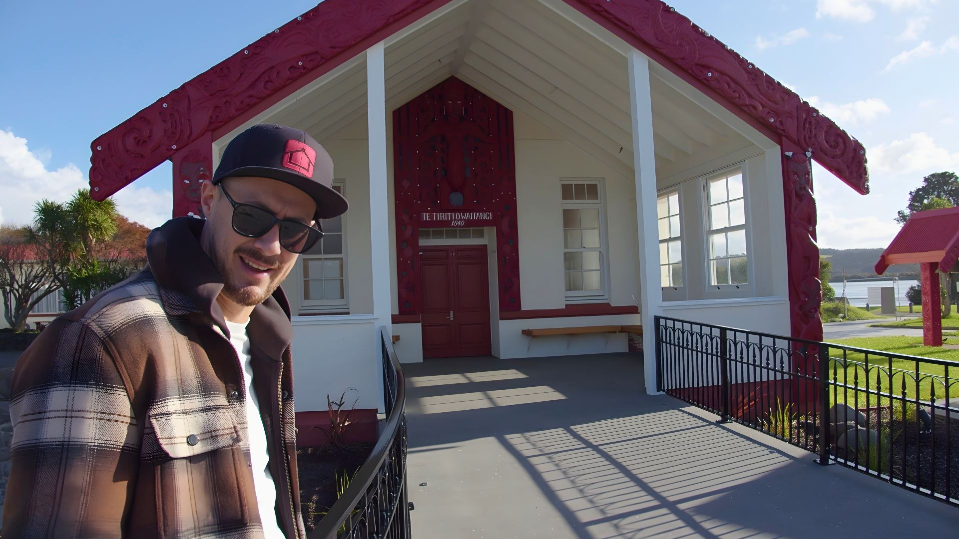 Man posing in front of a red and white marae with a red roof.