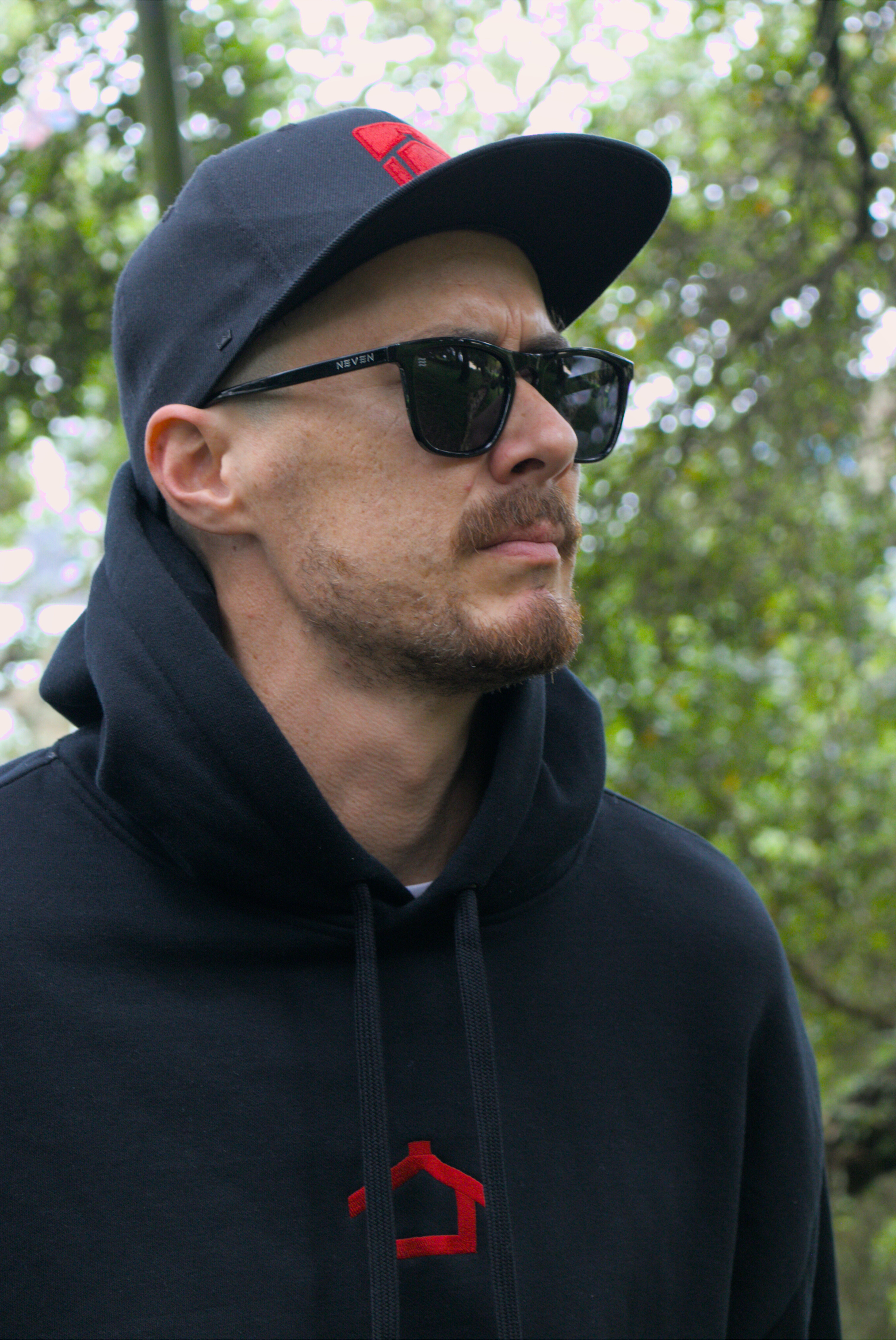 Man wearing a black hoodie and cap with a red logo, standing outdoors with greenery in the background.